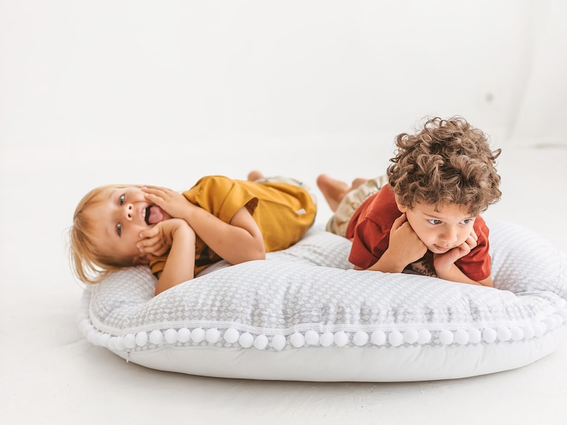 May include: Two children are lying on a white and gray round pillow with a white pom-pom trim. The child on the left is wearing a yellow shirt and has their hand on their cheek. The child on the right is wearing a red shirt and has their chin resting on their hands.