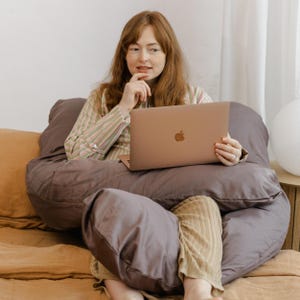 Puede incluir: Una persona sentada en una cama usa una computadora portátil, con una almohada corporal gris grande. Viste una camisa a rayas y pantalones color beige. La cama tiene una combinación de colores beige y marrón, con una lámpara blanca al fondo.
