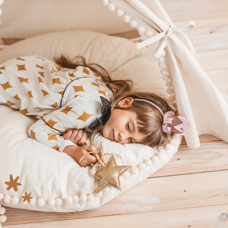 May include: A young girl sleeps peacefully in a white teepee tent, wearing a pink and gold crown headband. She is lying on a white and gold patterned pillow with a gold star-shaped toy.