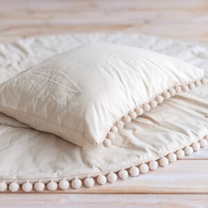 May include: A round, cream-colored play mat with a white pom-pom trim. A white pillow is placed on top of the mat.