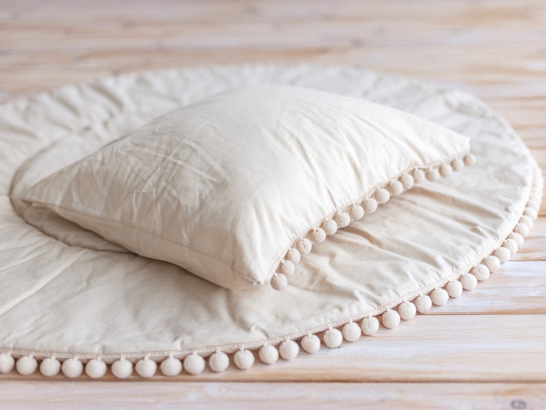 May include: A round, cream-colored play mat with a white pom-pom trim. A white pillow is placed on top of the mat.