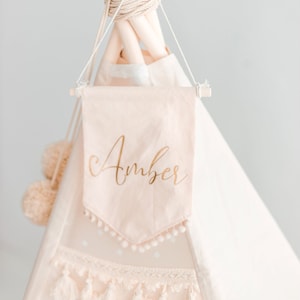 Può includere: Uno stendardo di tessuto rosa con il nome "Amber" scritto in lettere dorate. Lo stendardo è appeso a un teepee bianco con pompon bianchi.