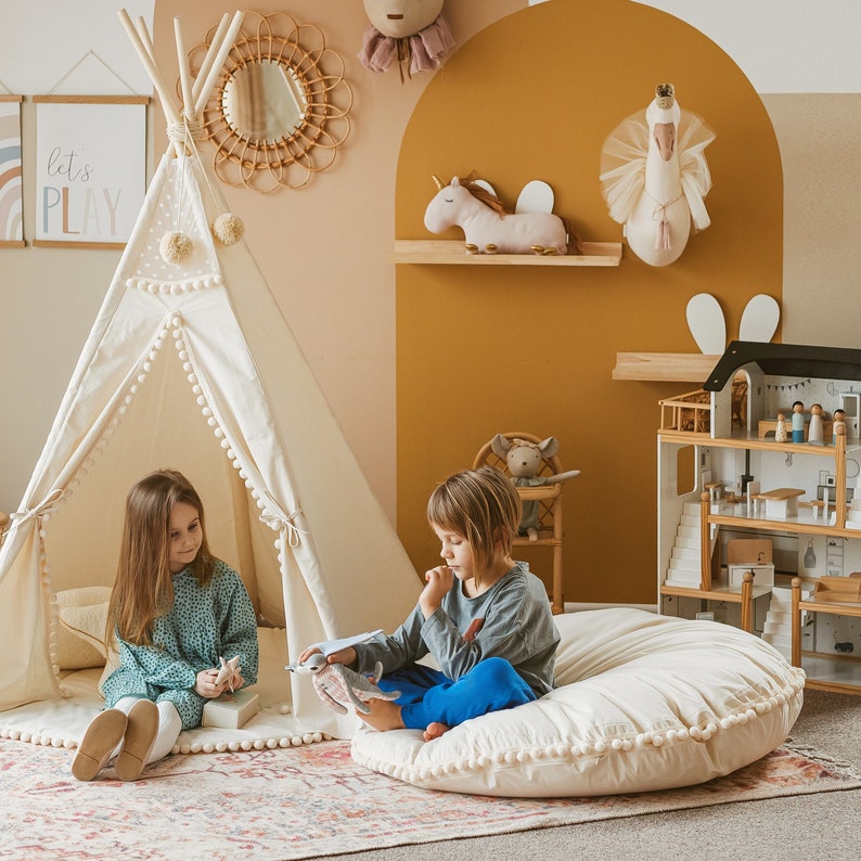 May include: Two children play in a room decorated with a teepee tent, a dollhouse, and stuffed animals. The teepee tent is white with a brown trim and has a white pom-pom trim. The dollhouse is made of wood and has a white roof. The stuffed animals include a unicorn, a swan, and a rabbit. The room is painted in a warm yellow color.