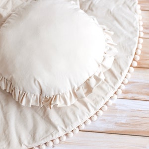 May include: A cream-colored round pillow with a ruffled edge sits on a cream-colored round rug with a white pom-pom trim. The rug is on a light wood floor.