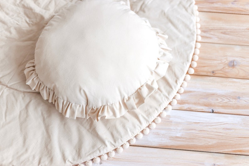 May include: A cream-colored round pillow with a ruffled edge sits on a cream-colored round rug with a white pom-pom trim. The rug is on a light wood floor.