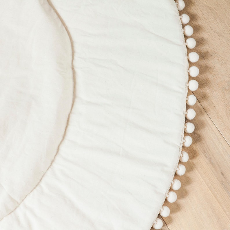 May include: A white round play mat with a white pom-pom trim around the edge.
