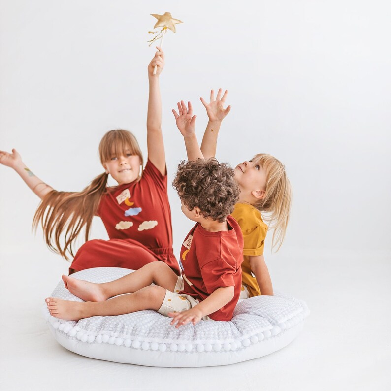 May include: Three children are playing on a white fluffy pillow. One child is holding a gold star wand. The child in the middle is wearing a red and orange shirt with a cloud and moon design. The child on the right is wearing a yellow shirt.