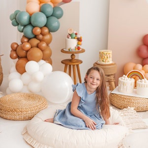 May include: A white, round floor cushion with a ruffled edge, measuring 40 inches in diameter. A young girl in a light blue dress is sitting on the cushion. The cushion is surrounded by a variety of colorful balloons, cakes, and other decorations.