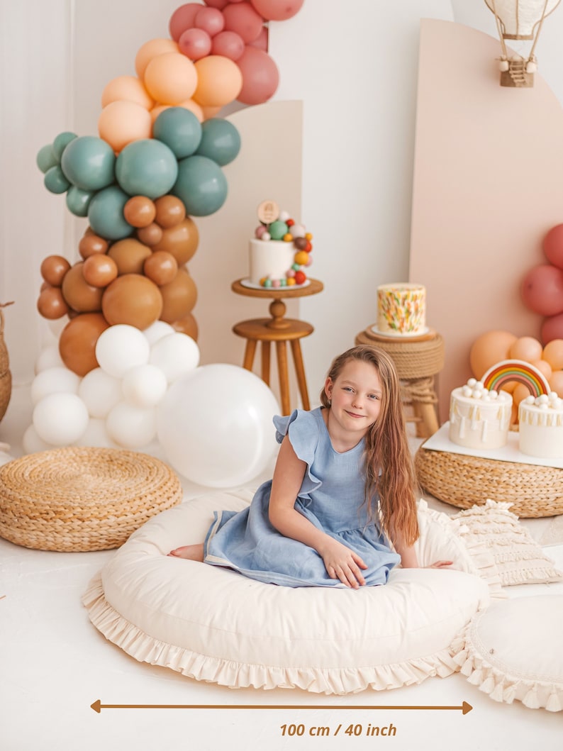 May include: A white, round floor cushion with a ruffled edge, measuring 40 inches in diameter. A young girl in a light blue dress is sitting on the cushion. The cushion is surrounded by a variety of colorful balloons, cakes, and other decorations.