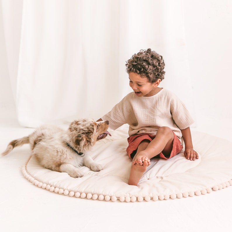 May include: A young child sits on a cream-colored play mat with a brown and white puppy. The mat has a white pom-pom trim. The child is wearing a cream-colored shirt and red shorts. The child is smiling and looking at the puppy.