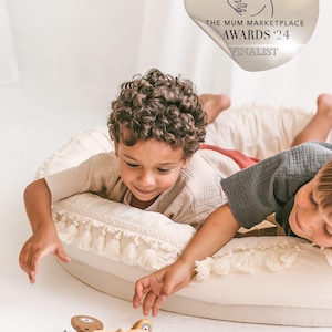 May include: Two children play with a wooden toy airplane on a cream-colored cushion with fringe. The toy airplane is pink and has a propeller. The children are wearing casual clothing. A sticker with the text "The Mum Marketplace Awards '24 Finalist" is visible in the background.