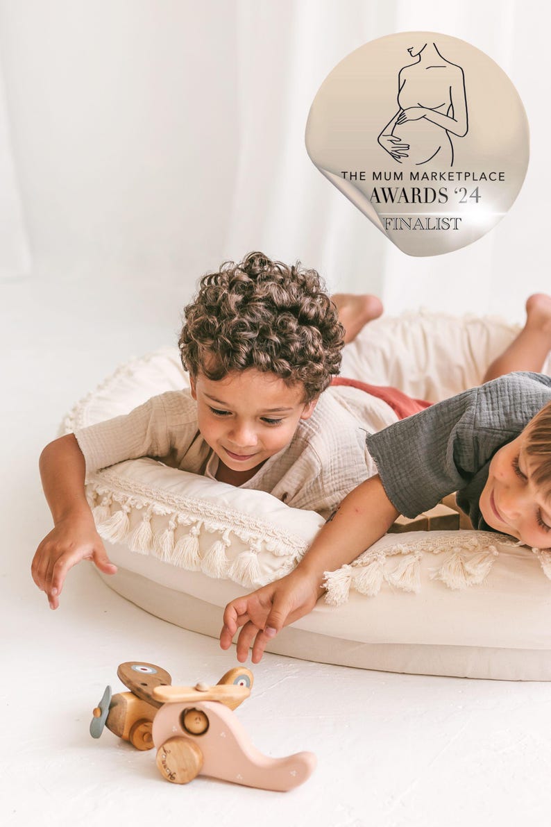 May include: Two children play with a wooden toy airplane on a cream-colored cushion with fringe. The toy airplane is pink and has a propeller. The children are wearing casual clothing. A sticker with the text "The Mum Marketplace Awards '24 Finalist" is visible in the background.