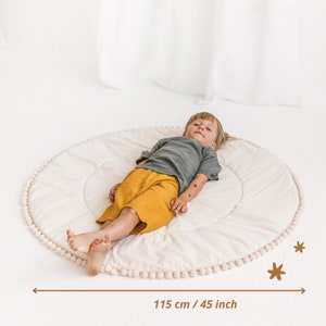 May include: A white round play mat with a white pom-pom trim. A child is lying on the mat. The mat measures 45 inches in diameter.