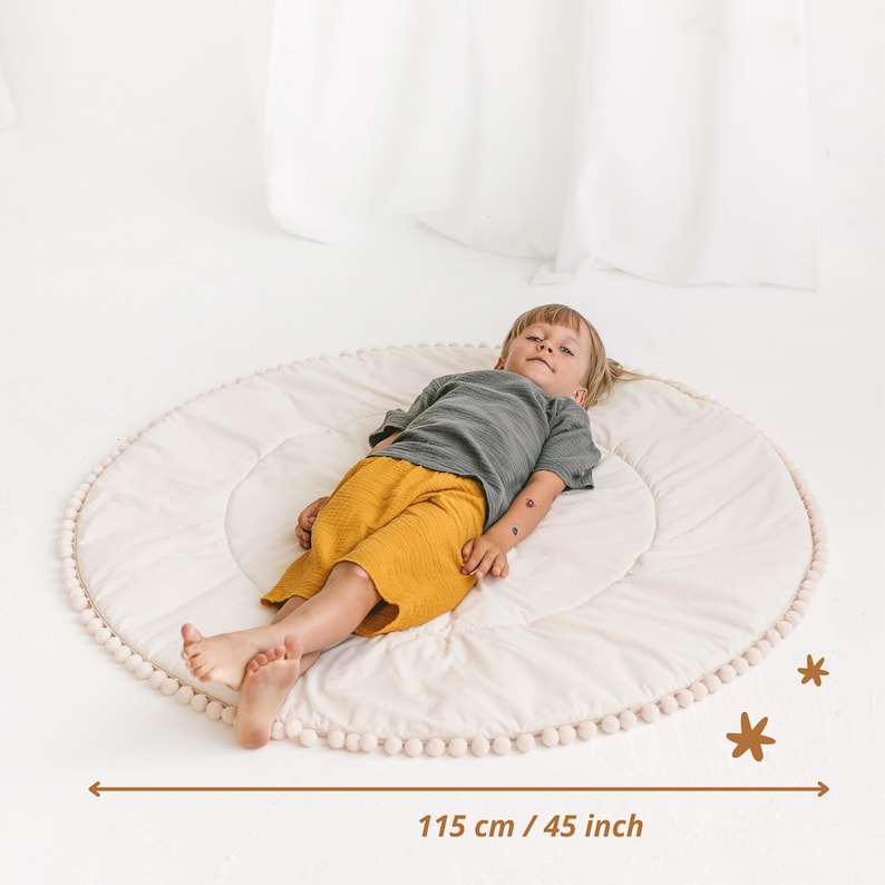 May include: A white round play mat with a white pom-pom trim. A child is lying on the mat. The mat measures 45 inches in diameter.