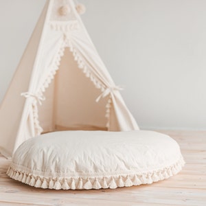 May include: A white round cushion with a fringe trim sits in front of a white teepee tent with a fringe trim. The cushion and teepee are on a light wood floor.