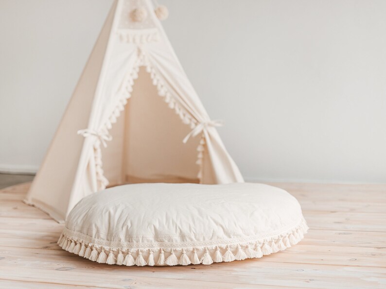May include: A white round cushion with a fringe trim sits in front of a white teepee tent with a fringe trim. The cushion and teepee are on a light wood floor.