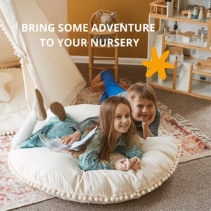 May include: Two children are sitting on a large, white, round cushion with a white pom-pom trim. The cushion is on a patterned rug. The girl is wearing a blue and green dress and the boy is wearing a blue shirt and jeans. The text "Bring some adventure to your nursery" is visible in the background.