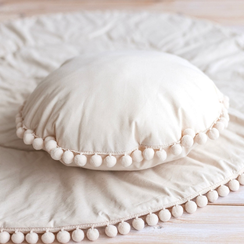 May include: A white round pillow with a white pom-pom trim. The pillow is sitting on a white fabric with a white pom-pom trim.