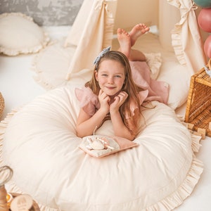May include: A young girl wearing a pink dress and a blue bow in her hair is sitting on a large, round, cream-colored pillow with a ruffle trim. She is holding a small, cream-colored pillow with a brown and white striped pattern. The pillow has a small, embroidered animal on it.