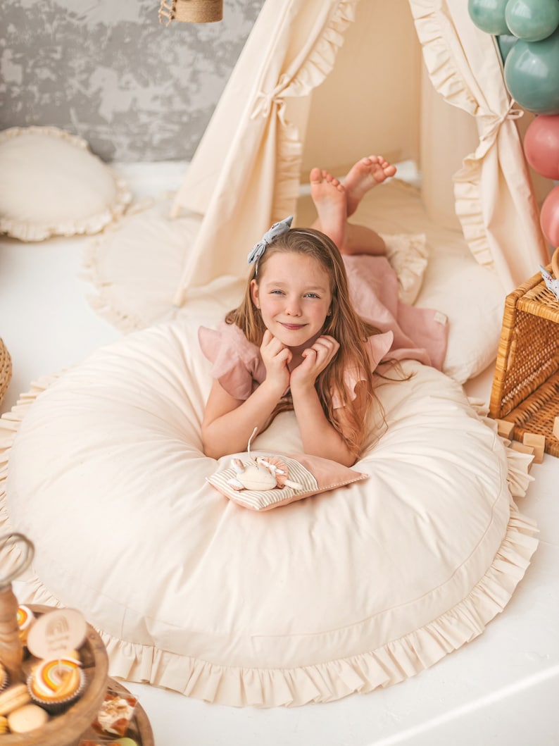 May include: A young girl wearing a pink dress and a blue bow in her hair is sitting on a large, round, cream-colored pillow with a ruffle trim. She is holding a small, cream-colored pillow with a brown and white striped pattern. The pillow has a small, embroidered animal on it.
