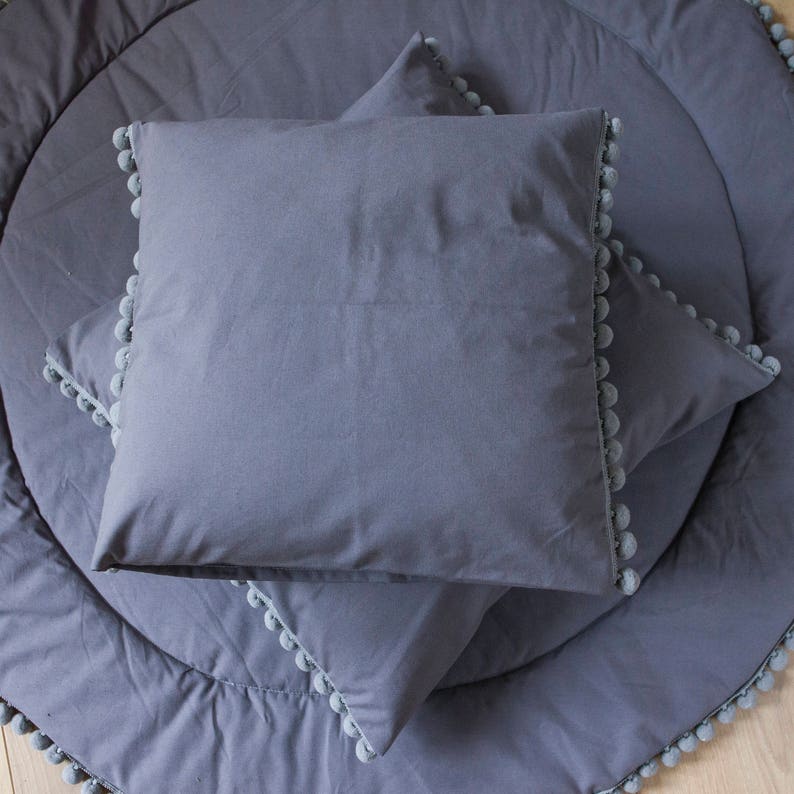 May include: Three gray throw pillows with pom-pom trim. The pillows are arranged on a gray round rug with pom-pom trim.