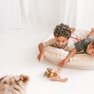 May include: Two children are lying on a white fringed cushion, reaching for a wooden toy airplane with pink and brown accents. The airplane has a propeller and wheels.