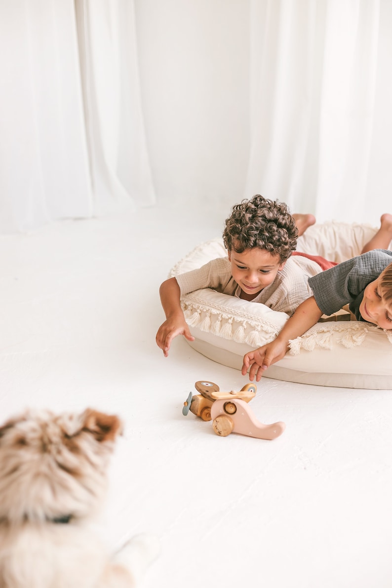 May include: Two children are lying on a white fringed cushion, reaching for a wooden toy airplane with pink and brown accents. The airplane has a propeller and wheels.