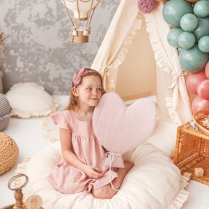 May include: A young girl sits on a cream-colored, ruffled floor cushion holding a pink heart-shaped balloon. She is wearing a pink dress and a pink headband. The background features a white teepee tent decorated with balloons and a white hot air balloon hanging from the ceiling.