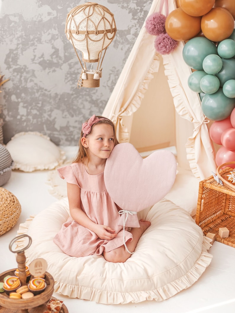 May include: A young girl sits on a cream-colored, ruffled floor cushion holding a pink heart-shaped balloon. She is wearing a pink dress and a pink headband. The background features a white teepee tent decorated with balloons and a white hot air balloon hanging from the ceiling.