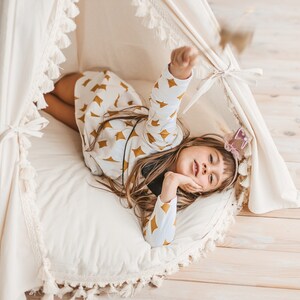 May include: A young girl wearing a white and gold patterned shirt lies in a white teepee with a white pillow and fringe trim. She is holding a sparkly wand and looking at the camera.