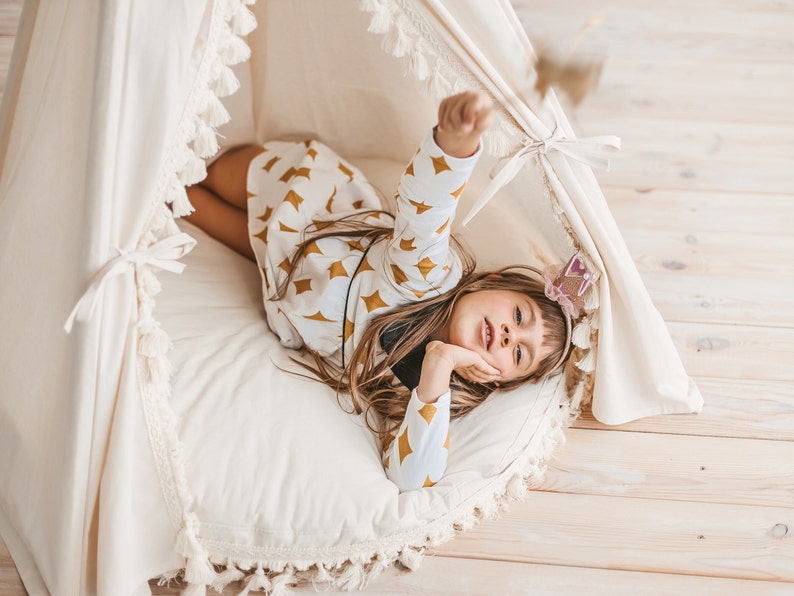 May include: A young girl wearing a white and gold patterned shirt lies in a white teepee with a white pillow and fringe trim. She is holding a sparkly wand and looking at the camera.