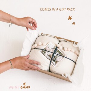 May include: A gift box with a white fabric inside, tied with a black ribbon and decorated with white pom-poms. The box is open and the fabric is partially pulled out. The box is on a white surface. The text "COMES IN A GIFT PACK" is written above the box.