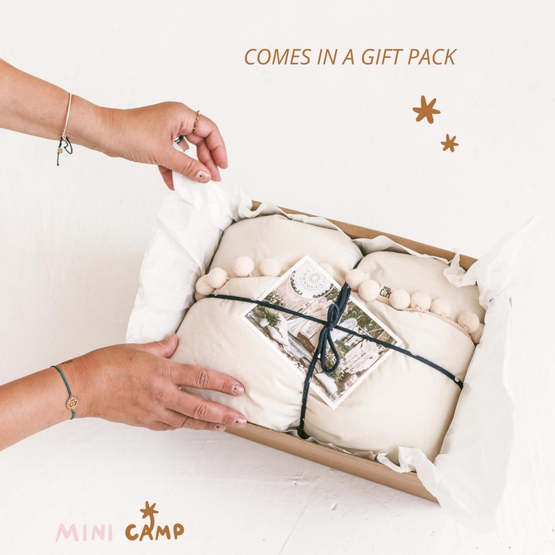 May include: A gift box with a white fabric inside, tied with a black ribbon and decorated with white pom-poms. The box is open and the fabric is partially pulled out. The box is on a white surface. The text "COMES IN A GIFT PACK" is written above the box.