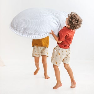 May include: Two children play with a large, round, white and gray play mat with a white pom-pom trim. The mat is being held up by one child, while the other child stands behind the mat.