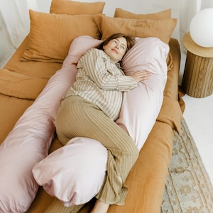 May include: A person lies on a bed with a light pink U-shaped body pillow. The bed has a rust-coloured duvet cover and pillows. The person wears a striped shirt and tan trousers. A small wooden side table with a white lamp is visible.