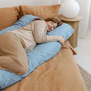 May include: A light blue U-shaped body pillow on a bed with tan bedding. The person is wearing a striped shirt and tan pants. A white sphere lamp sits on a small wooden side table.