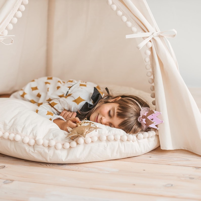 May include: A young girl wearing a crown sleeps on a white pillow with a gold star-shaped toy. The pillow has a white pom-pom trim.