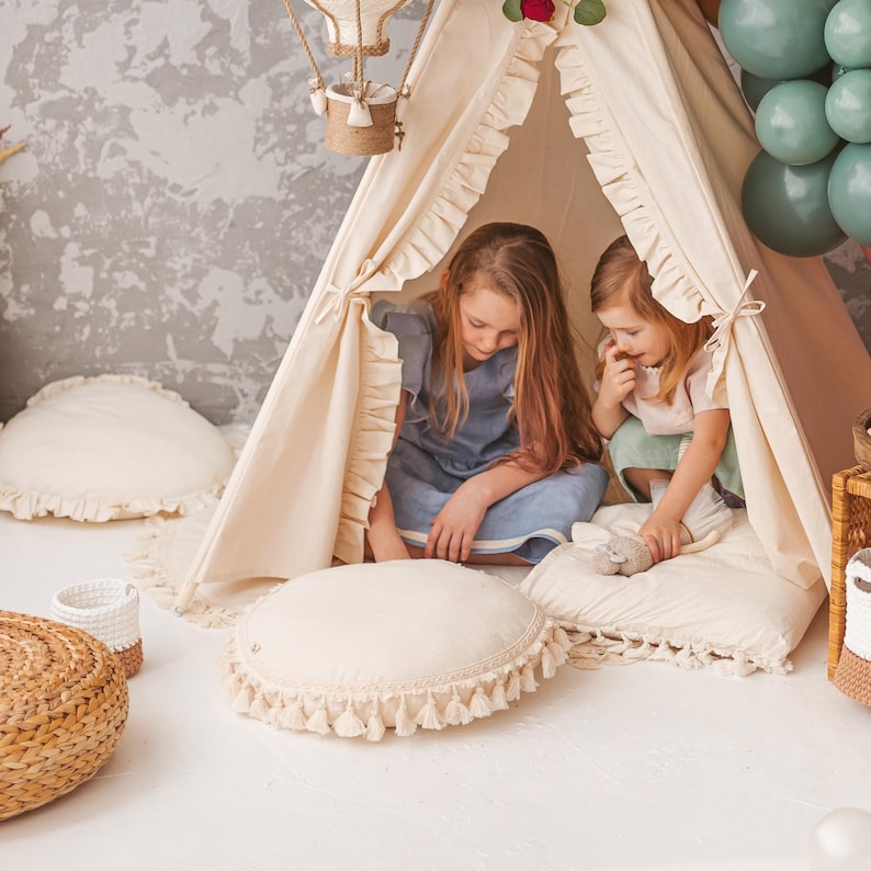 Op de afbeelding: Twee kinderen zitten in een witte tipi-tent met gekrulde randen. De tipi is versierd met een luchtballon en een rode bloem. De kinderen dragen casual kleding en kijken naar iets op hun schoot.