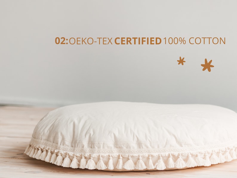 May include: A large, round, white cushion with a white fringe trim and tassels. The cushion is made of 100% cotton and is OEKO-TEX certified. The text "02: OEKO-TEX CERTIFIED 100% COTTON" is visible above the cushion.