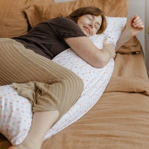 May include: A white body pillow with a floral pattern is being embraced. The pillow has a soft, curved shape, designed for side sleepers. The pillow is on a bed with brown bedding.