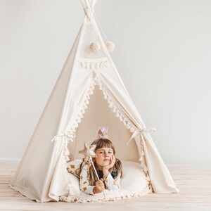 May include: A young girl wearing a crown and holding a star-shaped wand sits inside a white teepee with a fringe trim. The teepee is decorated with small white pom-poms.