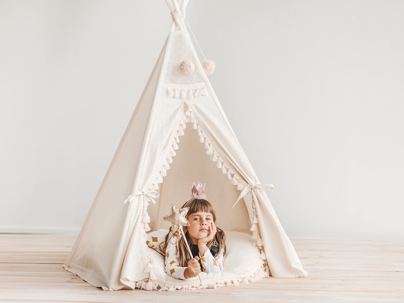 May include: A young girl wearing a crown and holding a star-shaped wand sits inside a white teepee with a fringe trim. The teepee is decorated with small white pom-poms.