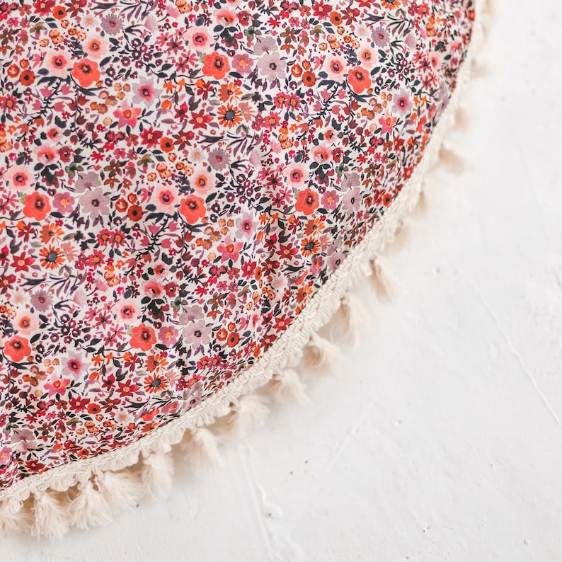 Pu&ograve; includere: Un cuscino rotondo con motivo floreale e bordo a frangia bianca. Il cuscino ha uno sfondo bianco con fiori rossi, rosa e arancioni.
