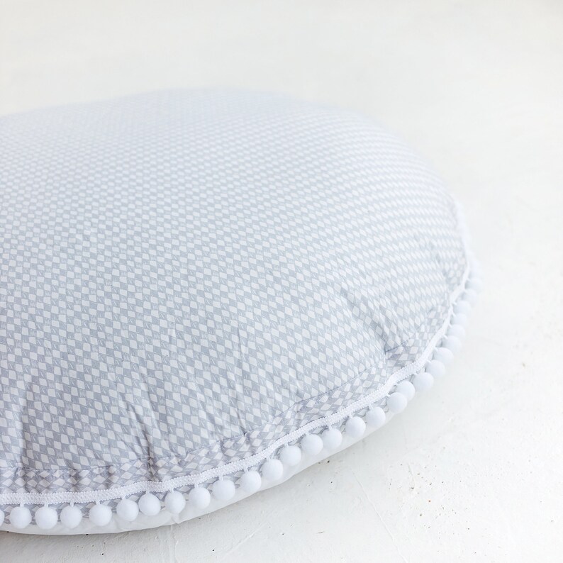 May include: A gray and white patterned floor cushion with a white pom-pom trim.