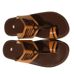 Könnte beinhalten: Ein Paar braune Ledersandalen mit Zehenriemen-Design. Die Sandalen haben einen hellbraunen Akzent am oberen Riemen und ein dunkelbraunes geflochtenes Detail. Das Fußbett ist in einem dunkleren Braunton gehalten, mit weißen Nähten an den Rändern.