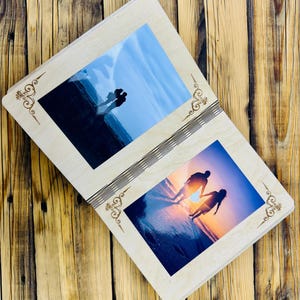 May include: A wooden photo album with two open pages, each displaying a photograph of a couple silhouetted against a sunset on a beach. The album has a decorative design on the front and back covers.