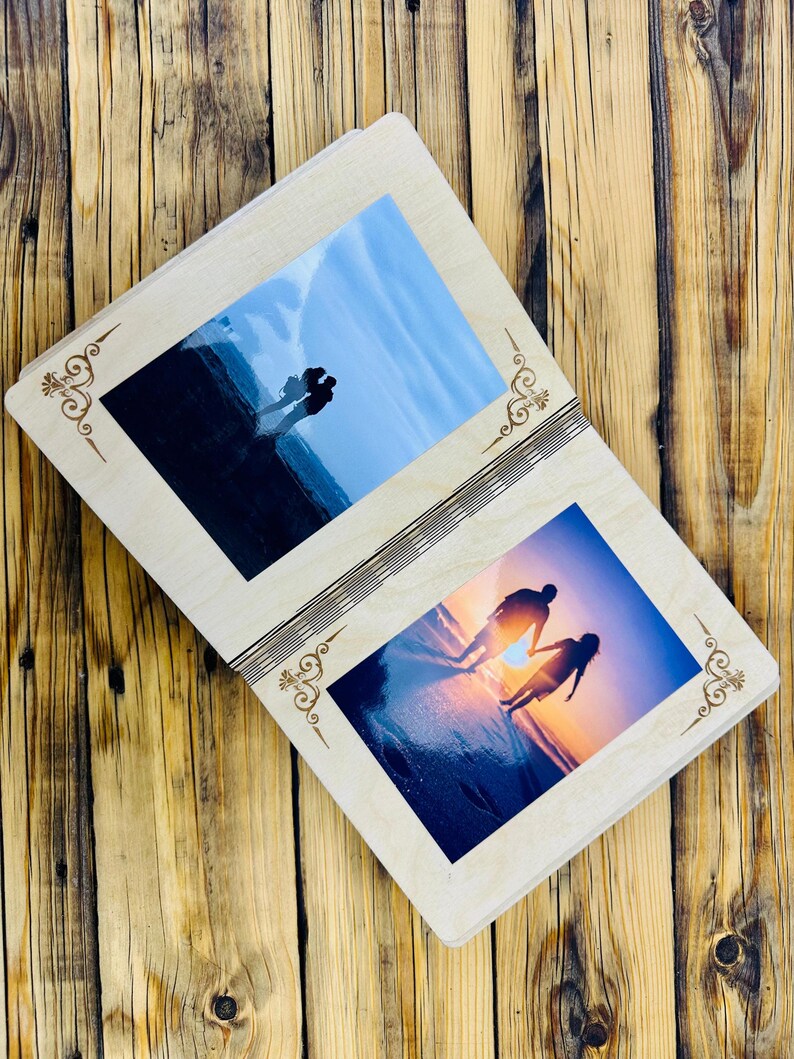 May include: A wooden photo album with two open pages, each displaying a photograph of a couple silhouetted against a sunset on a beach. The album has a decorative design on the front and back covers.