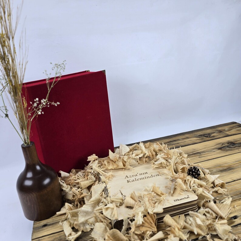 May include: A stack of wooden coasters with the text "Azra'un Kaleminden" in brown lettering on a light brown background. The coasters are surrounded by wood shavings and a red velvet box.