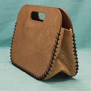 Op de afbeelding: Een bruine houten handtas met een zwarte gestikte rand. De tas heeft een bovenste handvat en een klep sluiting.