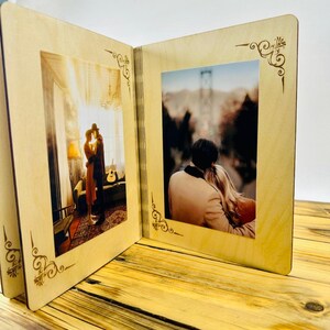 May include: A wooden photo album with two open pages, each displaying a photograph of a couple. The album has a light brown wood finish and intricate decorative designs around the edges of the photo frames.